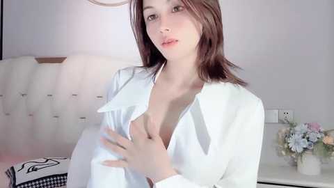 Media: Video of a young Caucasian woman with light skin and brown hair, wearing an unbuttoned white shirt, revealing cleavage, in a minimalist bedroom with a white headboard and floral arrangement on a nightstand.