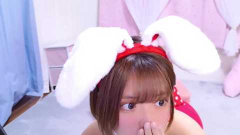 Media: Video of an Asian woman with short brown hair, wearing bunny ears and a red dress, kneeling on a white floor in a pink room.