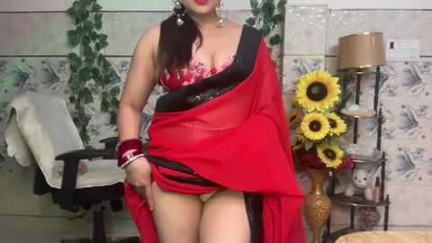 Media: Video of a voluptuous South Asian woman with dark hair, wearing a red sari, black bra, and gold jewelry, standing in a tiled room with floral wallpaper, greenery, and a sunflower vase.