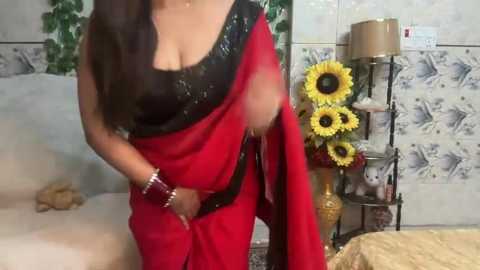 Media: Video of a South Asian woman in a red sari with black sequined blouse, gold bangles, holding sari, indoor setting with tiled walls, sunflowers, and a lamp.