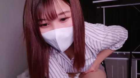 Media: Video of an East Asian woman with long, straight brown hair and light skin, wearing a white surgical mask, striped shirt, and black shorts. She leans forward, looking directly at the camera. Background shows a gray wall and a black curtain.