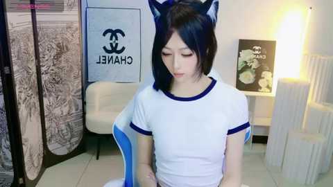 Media: Video of an East Asian woman in a white t-shirt with blue trim, wearing cat ears, sitting indoors with a white couch and framed art in the background.