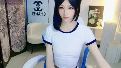 Media: Video of an East Asian woman with fair skin and straight black hair, wearing a white crop top with blue trim, sitting in a modern room with a white chair and Chanel posters.