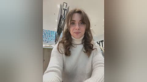 Media: Video of a young Caucasian woman with wavy brown hair, fair skin, and a slight smile, wearing a cream-colored ribbed sweater. The background features a modern, minimalist office with white walls, a black metal chair, and a blue box of documents.