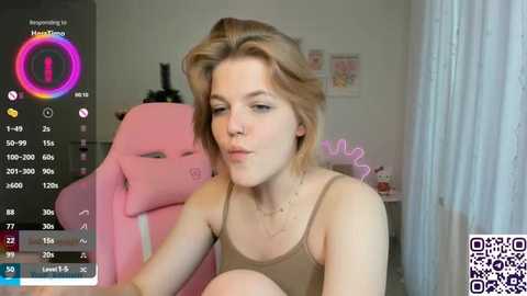 A video of a young woman with light skin and short blonde hair, wearing a brown tank top, sitting in a pink gaming chair in a cozy room with white curtains.