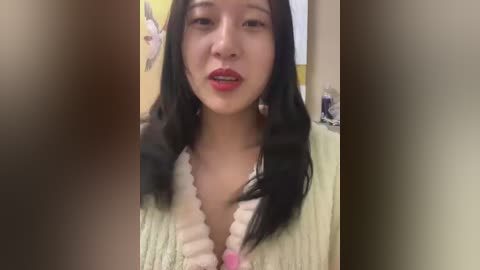 Media: Video of a young Asian woman with long black hair, wearing a cream-colored, ribbed sweater. She has a neutral expression with slightly parted lips and a subtle blush on her cheeks.