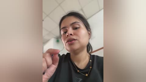 Media: Video of a young woman with medium brown skin and dark hair, wearing a black shirt and necklace, applying makeup in a room with a white ceiling and beige walls.