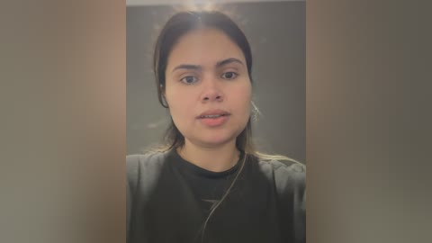 Media: Video of a young woman with olive skin, dark hair, and medium build, wearing a black t-shirt and dark hairnet, standing indoors against a blurred, neutral-colored background.