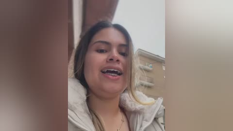 Media: Video of a young Latina woman with light brown skin and long blonde hair, wearing a beige jacket, smiling and looking up from a bathroom sink.