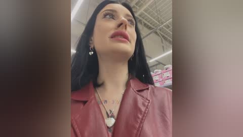 Media: Video of a woman with long black hair, wearing a red leather jacket and a silver necklace, standing in a brightly lit room with exposed ceiling beams.