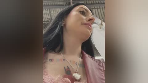 Media: Video of a woman with long black hair, light skin, and tattoos on her chest, wearing a pink lace top and a silver necklace with a heart pendant. Background shows a blurred, industrial setting with metal racks.