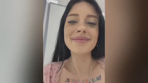 Media: Video of a smiling young woman with long black hair, light skin, and a pink floral top. She's indoors, with a blurred background.