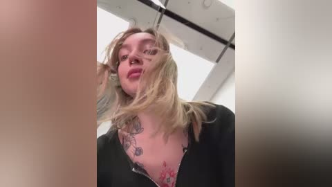 Media: Video of a blonde woman with a tattoo on her chest, wearing a black top, captured from a low angle, slightly blurred, in a modern interior with white walls and a circular skylight.