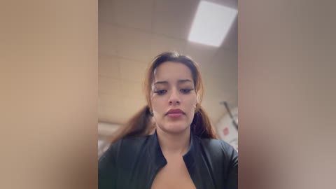 A video of a young woman with light brown skin, straight brown hair, and full lips, wearing a black jacket, taken in a dimly lit, institutional setting.