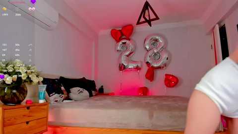 Media: Video of a bedroom with a bed, wooden dresser, and a vase of white flowers on the left. Silver balloons with \"28\" and red heart decorations hang on the white wall, illuminated by red light.