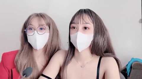 Media: Video of two young Asian women with long brown hair, wearing white surgical masks and round glasses, seated close together in a casual setting with neutral background.