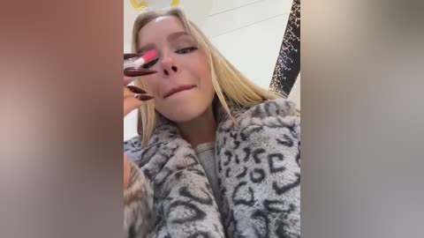 Video of a blonde woman in a leopard-print robe, applying red lipstick with a brush. The background features a tiled wall and a patterned curtain.