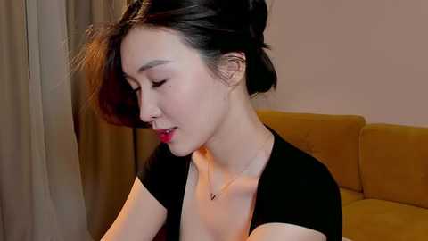 Media: A video of an East Asian woman with fair skin, dark hair in a messy bun, wearing a black top, looking down with a contemplative expression. Background features a mustard-yellow couch and beige curtains.