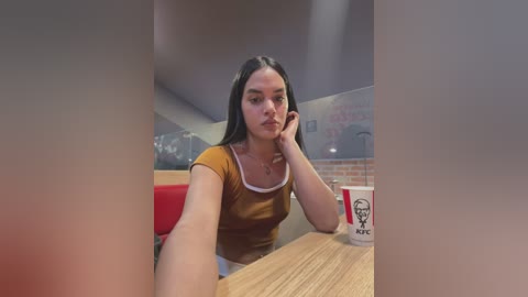 Media: Video of a young Latina woman with long black hair, wearing a mustard-colored top, resting her chin on her hand at a fast-food restaurant table. Background features a red chair, white coffee cup, and brick wall.