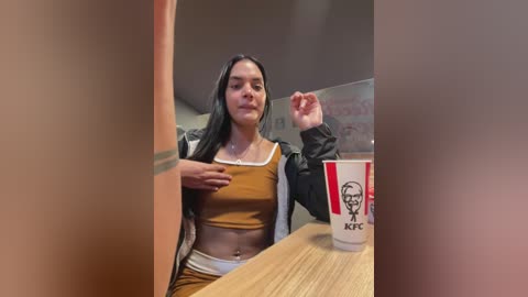 Media: Video of a Latina woman with long black hair, wearing a mustard crop top and black jacket, sitting at a wooden table with a KFC cup, in a fast-food restaurant.