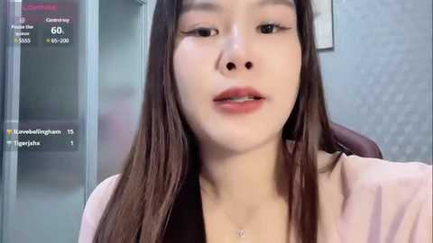 Media: Video of a young East Asian woman with long, straight, dark brown hair, wearing a pink cardigan, sitting in an office with a gray wall and a monitor displaying her profile details.