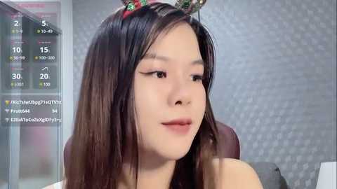 Media: Video of an Asian woman with long, straight black hair, wearing a red and green Christmas headband, in a modern, grey-brick-wall room, with a live stream overlay.