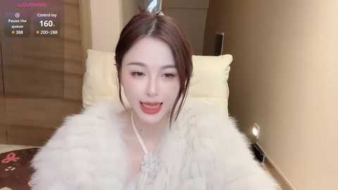 Media: Video of a young Asian woman with fair skin, brown hair, and light makeup, wearing a fluffy white fur coat, smiling while sitting on a beige bed with cream pillows in a cozy, warmly lit room.