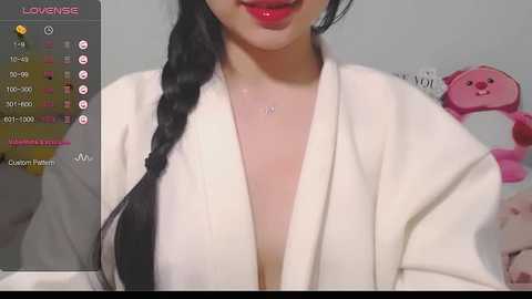 Media: Video of an Asian woman with a fair complexion, wearing a white robe with a deep V-neck, showcasing cleavage. Her long, braided black hair falls over her shoulder. The background features a pink teddy bear and a grey wall.