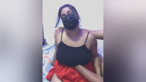 Media: A video of a young woman with light brown skin, wearing a black face mask and a black tank top with red skirt, posing indoors against a blurred background.