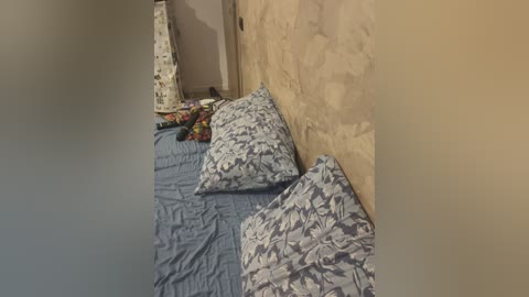 Media: Video of a rustic bedroom corner featuring a bed with blue and white patterned sheets, a beige stone wall, a white patterned lampshade, and scattered colorful clothes on the floor.