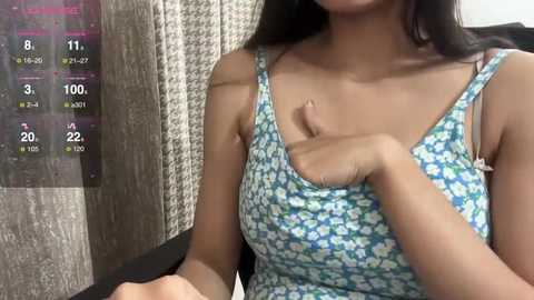 Media: Video of a young Asian woman with medium skin tone, wearing a blue floral-patterned tank top, sitting with her hand on her breast. Background features a grey textured wall and a digital screen showing health metrics.