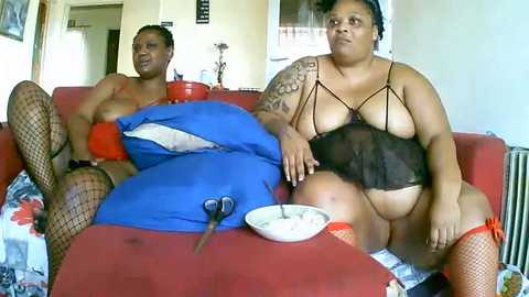 Media: Video of two plus-sized Black women in revealing lingerie, one with a tattoo, sitting on a red couch with a blue pillow.