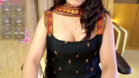 Media: Video of a woman in a black, sleeveless top adorned with gold embroidery, seated indoors with a warm-toned backdrop and digital display showing weather data.