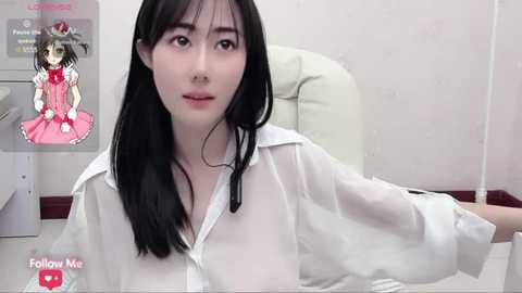 Media: Video of an East Asian woman with straight black hair, wearing a white blouse, sitting on a white chair. Background shows a poster of a character in a pink dress.