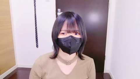 Media: Video of an East Asian woman with straight black hair and a black face mask, wearing a beige top and a silver necklace. She stands indoors, against a light wooden wall and dark wooden door, with a black-and-white striped tie hanging behind her.