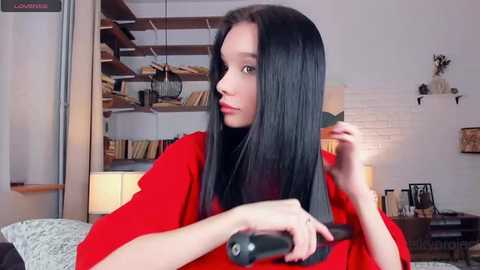 Media: Video of a young woman with long, straight black hair, sitting in a cozy living room, holding a hairdryer while looking in a mirror.