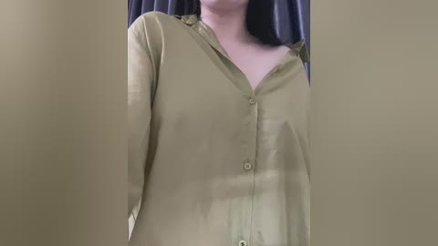 Media: Video of a person wearing a loose-fitting, olive-green button-down shirt with long sleeves. The background is a muted, olive-green wall, creating a monochromatic effect. The person\u2019s face is not visible, focusing on the upper torso.