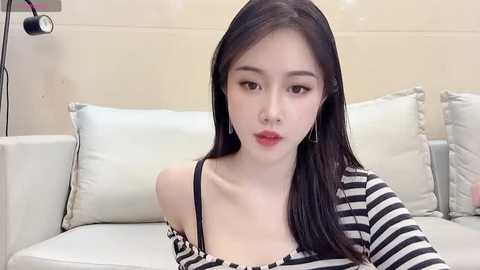 Media: Video of an East Asian woman with fair skin, long black hair, and red lipstick, wearing a black and white striped off-shoulder top, sitting on a white couch in a modern room.