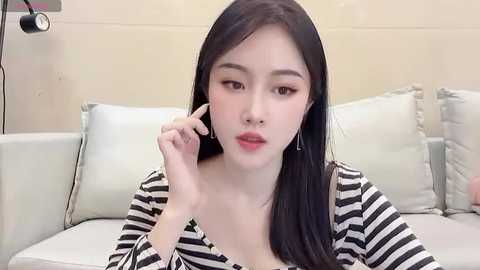 Media: Video of an East Asian woman with long black hair, fair skin, and red lipstick, wearing a black-and-white striped shirt, adjusting her earpiece on a white couch.