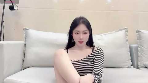 Media: Video of an East Asian woman with long black hair, wearing a black and white striped top, sitting on a white sofa.