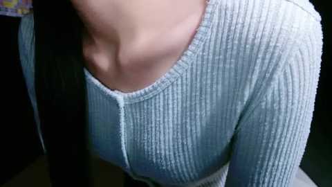 Media: A close-up video of a young woman's neck and chest, wearing a light blue, ribbed knit sweater. Her skin is light, and the background is blurred, highlighting her attire.