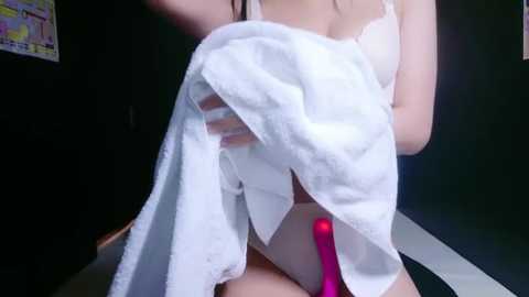 Media: A video of a woman holding a white towel against her groin, revealing a pink vibrator. She wears white lingerie. Dark wall background.