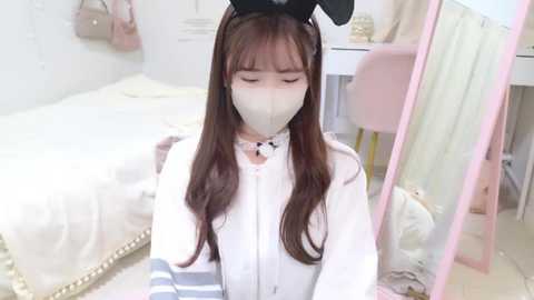 Media: Video of a young Asian woman with long brown hair, wearing a white dress, black bunny ears, and a white face mask, sitting on a bed in a softly lit, pastel-colored bedroom.