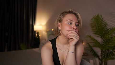 Media: Video of a blonde woman with a short bob haircut, wearing a black dress, thoughtfully resting her chin on her hand in a dimly lit, cozy indoor setting with a lit lamp, green plant, and dark curtains.