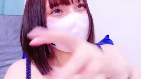 Media: Video of a young Asian woman with straight, shoulder-length dark brown hair, wearing a white surgical mask and a blue ribbon. She gestures with her right hand towards the camera.