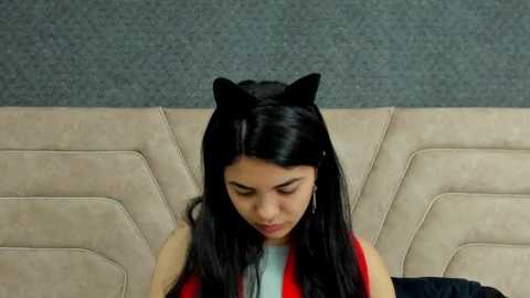 Media: Video of a young woman with long black hair and cat ears, wearing a sleeveless red dress, sitting on a beige leather couch with a gray carpeted background.