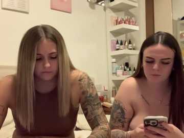 Media: Video of two tattooed women, one topless, sitting on a bed in a cluttered bedroom. The topless woman with large breasts holds a phone, while the other, wearing a brown top, looks down.