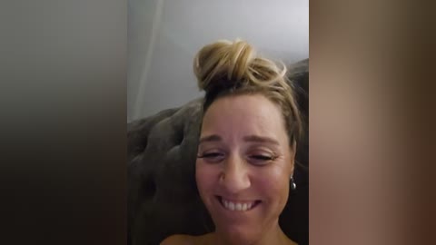 Media: Video of a Caucasian woman with light brown hair in a messy bun, smiling widely, with closed eyes, wearing a brown shirt. She's indoors on a dark couch, blurred background.