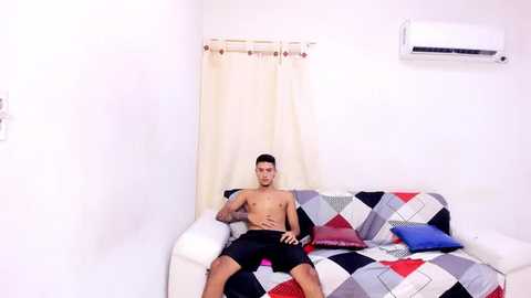 Media: Video of a muscular, shirtless man with short dark hair, wearing black shorts, reclining on a colorful quilted couch against a white wall with a cream curtain and air conditioner.