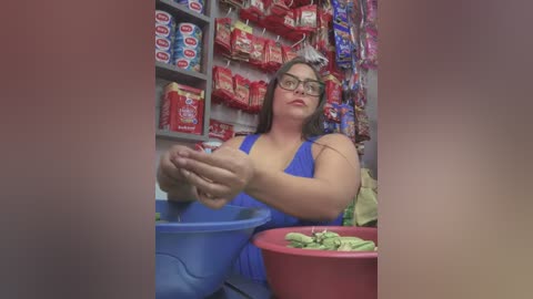 Media: Video of a plus-sized woman with glasses, wearing a blue tank top, weighing green vegetables in a market stall filled with red packets of snacks.